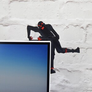 May include: Black silhouette of a zombie figure with red eyes, climbing on top of a computer monitor. The zombie is made of plastic and is a desk accessory.