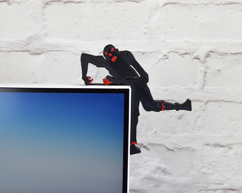 May include: Black silhouette of a zombie figure with red eyes, climbing on top of a computer monitor. The zombie is made of plastic and is a desk accessory.