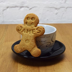 Puede incluir: Una galleta de jengibre con forma de persona con el nombre "JAMES" escrito en ella. La galleta est&aacute; sobre un platillo negro junto a una taza de caf&eacute; blanca con letras negras y grises.
