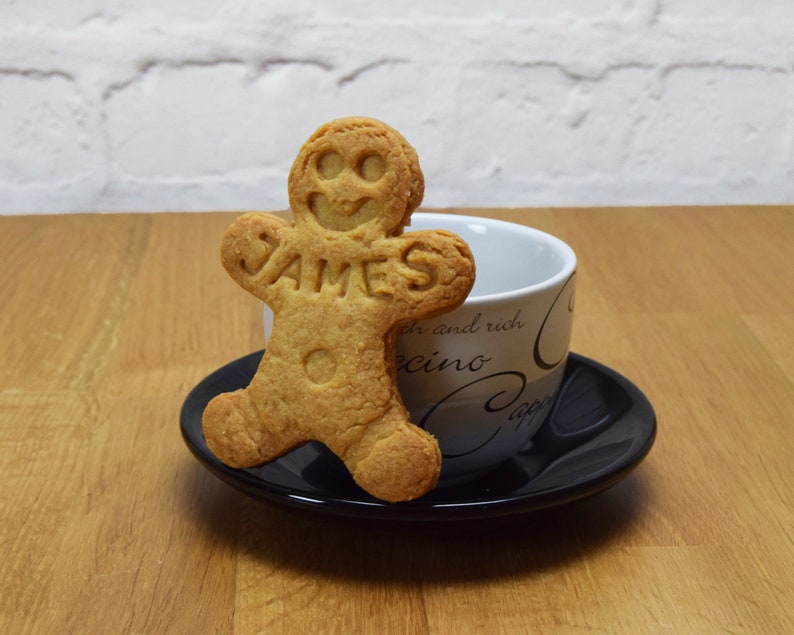 Puede incluir: Una galleta de jengibre con forma de persona con el nombre "JAMES" escrito en ella. La galleta est&aacute; sobre un platillo negro junto a una taza de caf&eacute; blanca con letras negras y grises.