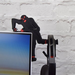 May include: A black 3D printed zombie figure with red eyes is perched on top of a computer monitor. The zombie is holding onto the monitor with its hands and has its legs dangling off the side. A pair of black headphones are hanging on the side of the monitor.