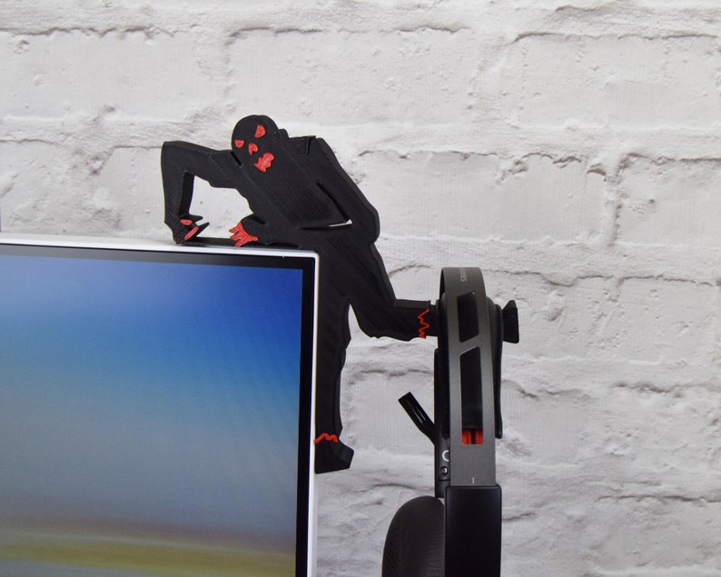 May include: A black 3D printed zombie figure with red eyes is perched on top of a computer monitor. The zombie is holding onto the monitor with its hands and has its legs dangling off the side. A pair of black headphones are hanging on the side of the monitor.