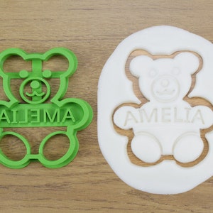 Puede incluir: Cortador de galletas de pl&aacute;stico verde en forma de oso de peluche con el nombre "AIJEMA" escrito en la parte delantera. Se muestra una masa de galletas blanca en el fondo con la misma forma de oso de peluche recortada y el nombre "AMELIA" escrito en la parte delantera.