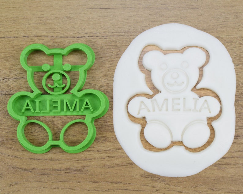 Puede incluir: Cortador de galletas de pl&aacute;stico verde en forma de oso de peluche con el nombre "AIJEMA" escrito en la parte delantera. Se muestra una masa de galletas blanca en el fondo con la misma forma de oso de peluche recortada y el nombre "AMELIA" escrito en la parte delantera.