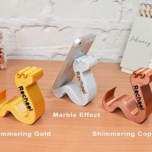 May include: Three 3D printed phone stands shaped like a worm with the name "Rachael" engraved on each. The stands are in three different finishes: shimmering gold, marble effect, and shimmering copper.