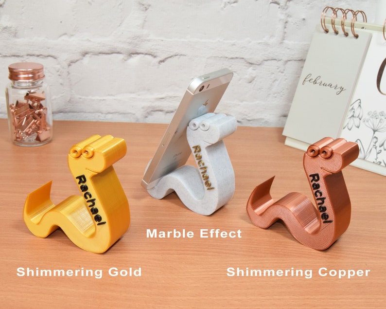 May include: Three 3D printed phone stands shaped like a worm with the name "Rachael" engraved on each. The stands are in three different finishes: shimmering gold, marble effect, and shimmering copper.