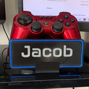 Pu&ograve; includere: Un controller per videogiochi rosso &egrave; seduto su un supporto nero con un bordo blu neon. Il supporto ha il nome "Jacob" scritto in lettere bianche su uno sfondo nero.