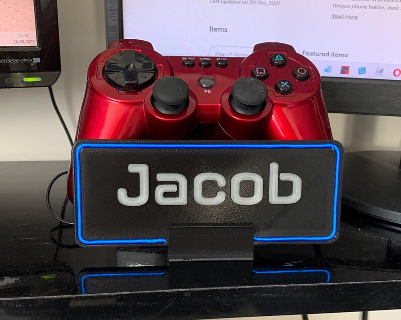 Pu&ograve; includere: Un controller per videogiochi rosso &egrave; seduto su un supporto nero con un bordo blu neon. Il supporto ha il nome "Jacob" scritto in lettere bianche su uno sfondo nero.