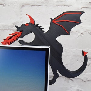May include: A black dragon-shaped corner shelf with red accents, including horns, eyes, and flames. The dragon is positioned on a white brick wall, with the shelf extending over the corner of a computer monitor.
