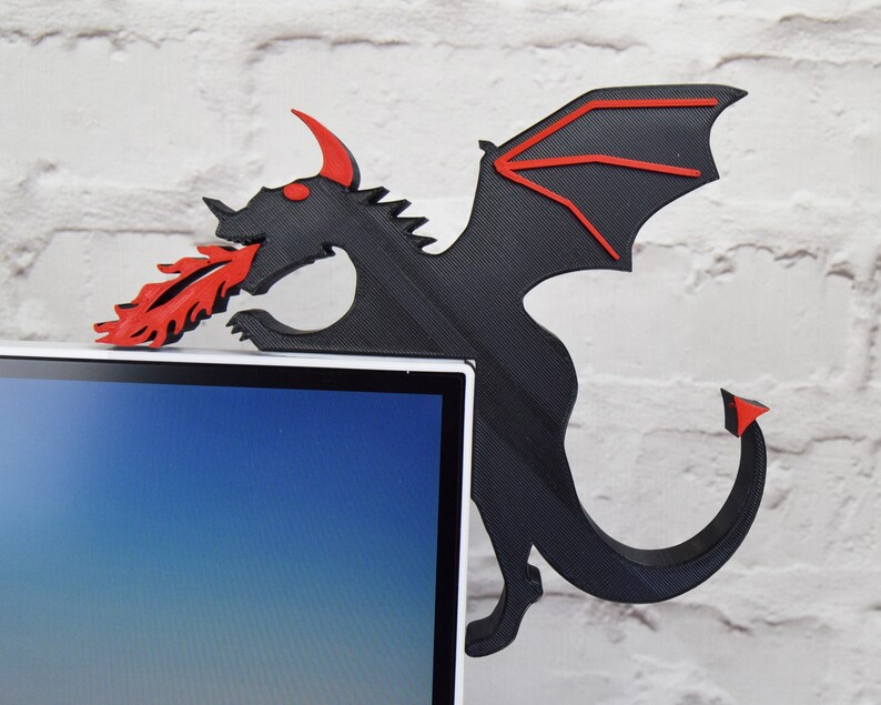 May include: A black dragon-shaped corner shelf with red accents, including horns, eyes, and flames. The dragon is positioned on a white brick wall, with the shelf extending over the corner of a computer monitor.