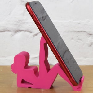 May include: A pink phone stand shaped like a person doing yoga. The stand is holding a red smartphone.