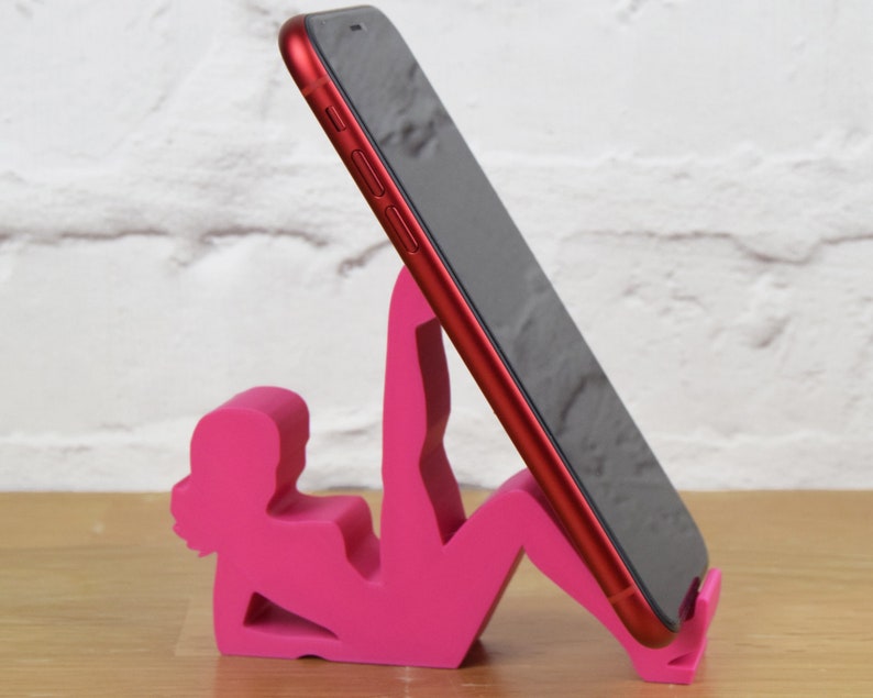 May include: A pink phone stand shaped like a person doing yoga. The stand is holding a red smartphone.