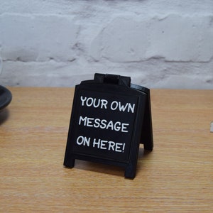 May include: A small, black A-frame chalkboard sign displaying the white text "YOUR OWN MESSAGE ON HERE!". The sign is on a wooden surface. A white mug and saucer are visible in the background. The sign is likely for displaying messages.