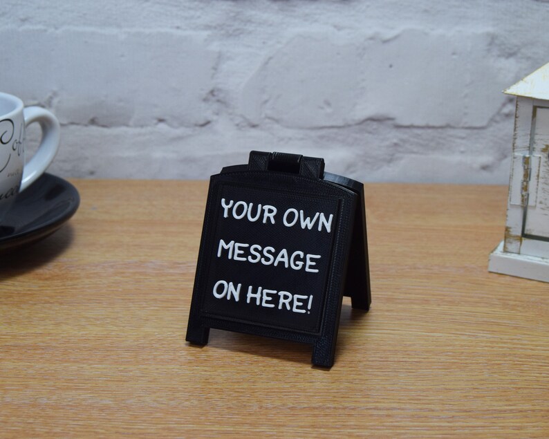 May include: A small, black A-frame chalkboard sign displaying the white text "YOUR OWN MESSAGE ON HERE!". The sign is on a wooden surface. A white mug and saucer are visible in the background. The sign is likely for displaying messages.