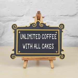 May include: Black sign with gold trim on a wooden easel. The sign reads "Unlimited Coffee with All Cakes".