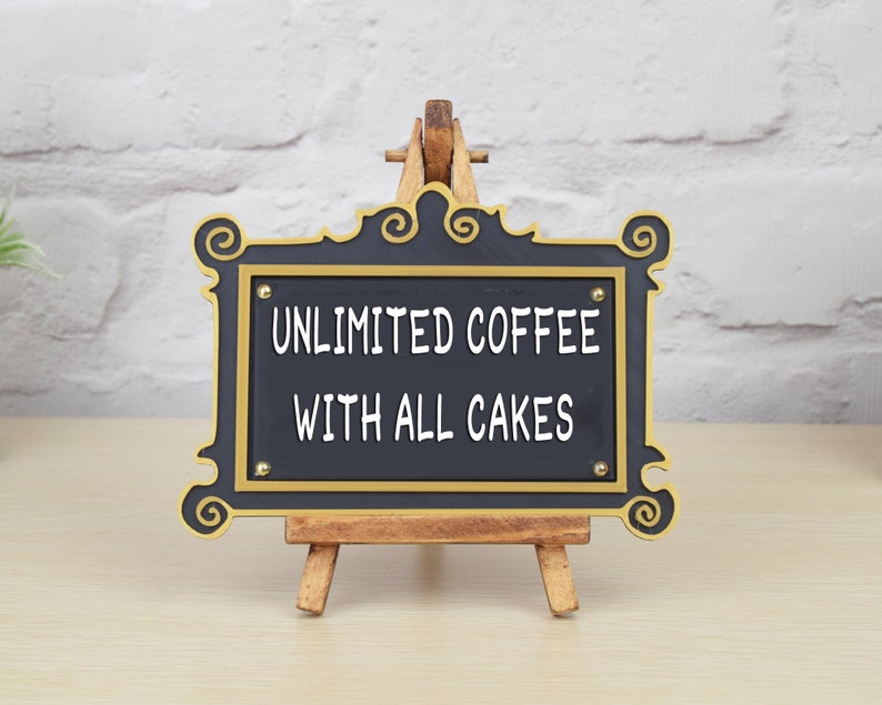 May include: Black sign with gold trim on a wooden easel. The sign reads "Unlimited Coffee with All Cakes".