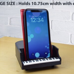 Puede incluir: Soporte para tel&eacute;fono negro con forma de piano con un dise&ntilde;o de teclado blanco. El soporte sostiene un tel&eacute;fono inteligente con una funda roja. La pantalla del tel&eacute;fono muestra la hora "12:34" y la fecha "domingo 14 de julio". El soporte est&aacute; dise&ntilde;ado para sostener tel&eacute;fonos de hasta 10,75 cm de ancho.