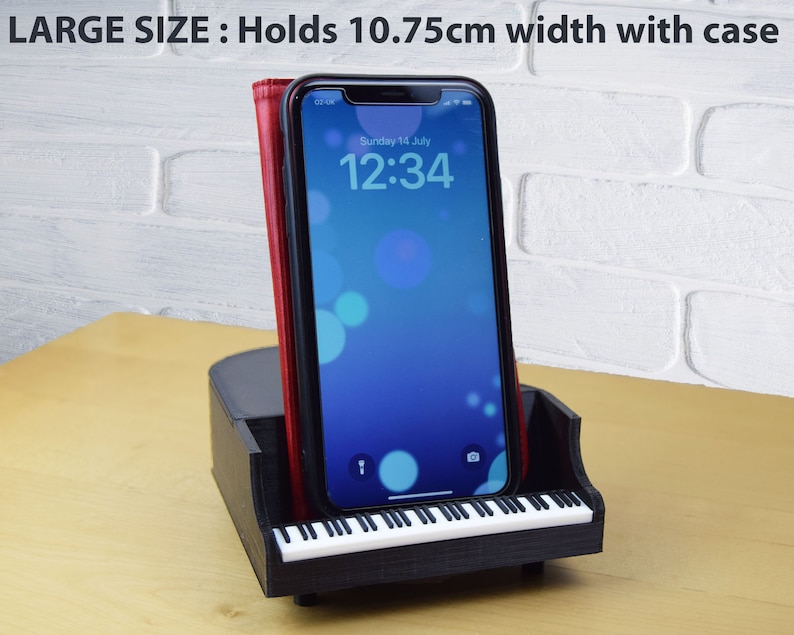 Puede incluir: Soporte para tel&eacute;fono negro con forma de piano con un dise&ntilde;o de teclado blanco. El soporte sostiene un tel&eacute;fono inteligente con una funda roja. La pantalla del tel&eacute;fono muestra la hora "12:34" y la fecha "domingo 14 de julio". El soporte est&aacute; dise&ntilde;ado para sostener tel&eacute;fonos de hasta 10,75 cm de ancho.