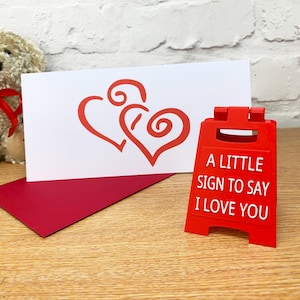 Op de afbeelding: Een rode kaart met twee rode harten en de tekst "A little sign to say I love you" op een witte achtergrond. De kaart ligt naast een rood, staand bord met dezelfde tekst.