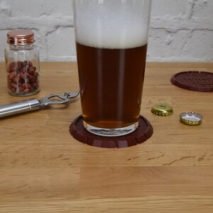 May include: A pint glass filled with beer sits on a brown coaster. A bottle opener, a jar of peanuts, and two bottle caps are on a wooden surface. Another coaster with the text "Your Words Here" is also visible.