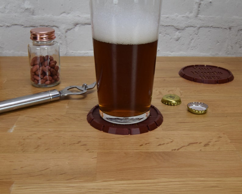 May include: A pint glass filled with beer sits on a brown coaster. A bottle opener, a jar of peanuts, and two bottle caps are on a wooden surface. Another coaster with the text "Your Words Here" is also visible.