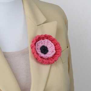 May include: A pink and red crocheted poppy flower brooch with a black center. The brooch is pinned to a beige blazer.