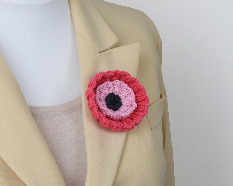 May include: A pink and red crocheted poppy flower brooch with a black center. The brooch is pinned to a beige blazer.