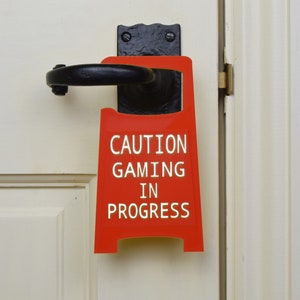 Op de afbeelding: Een rood deurschild met de tekst "CAUTION GAMING IN PROGRESS" in witte letters. Het bord hangt aan een witte deur met een zwarte deurknop.