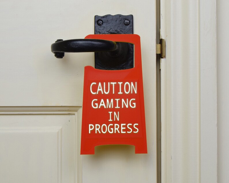 Op de afbeelding: Een rood deurschild met de tekst "CAUTION GAMING IN PROGRESS" in witte letters. Het bord hangt aan een witte deur met een zwarte deurknop.