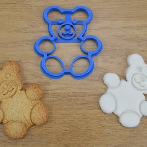 May include: Three bear-shaped cookie cutters, one blue plastic, one white, and one brown cookie. The blue plastic cookie cutter is on a wooden surface.
