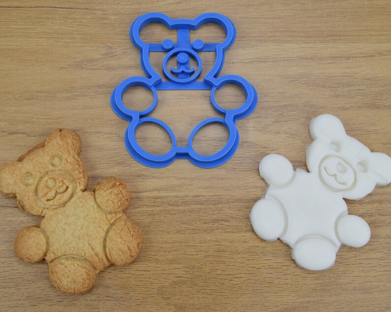 May include: Three bear-shaped cookie cutters, one blue plastic, one white, and one brown cookie. The blue plastic cookie cutter is on a wooden surface.