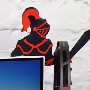 Op de afbeelding: Een zwart en rode 3D-geprinte koptelefoonstandaard in de vorm van een ridder. De ridder draagt een helm en een harnas. De standaard houdt een paar zwarte koptelefoons vast.