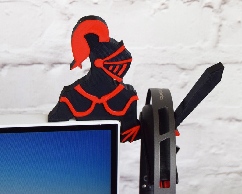 Op de afbeelding: Een zwart en rode 3D-geprinte koptelefoonstandaard in de vorm van een ridder. De ridder draagt een helm en een harnas. De standaard houdt een paar zwarte koptelefoons vast.