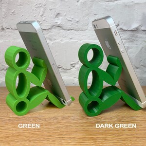May include: Two phone stands made of green plastic, shaped like a person sitting down. The stand on the left is a lighter shade of green, while the stand on the right is a darker shade of green.