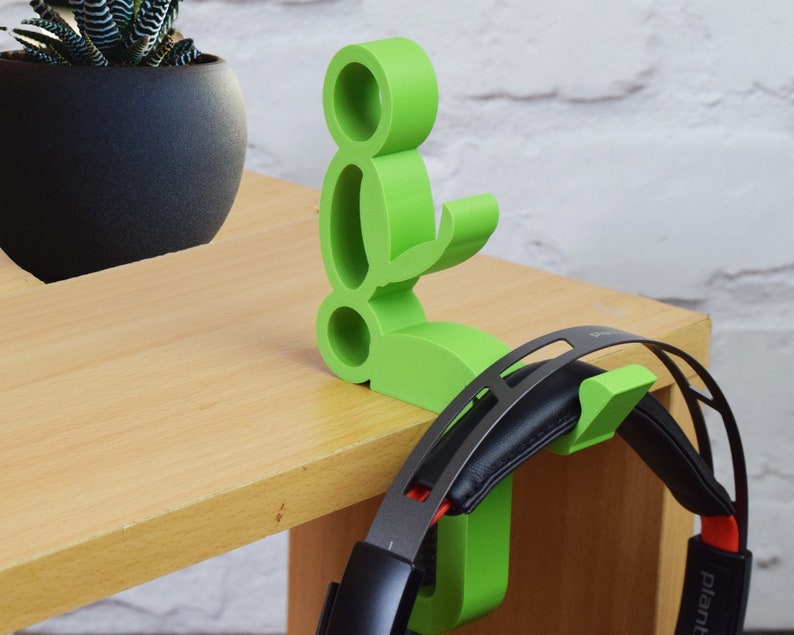 May include: A green 3D printed headphone stand shaped like a person sitting down. The stand has a hook on the back for hanging headphones. The stand is on a wooden table.