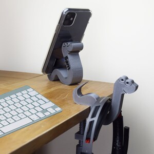 May include: Two 3D printed phone stands shaped like dinosaurs. The stand on the left is holding a black smartphone. The stand on the right is holding a pair of headphones. Both stands are on a wooden desk with a keyboard.