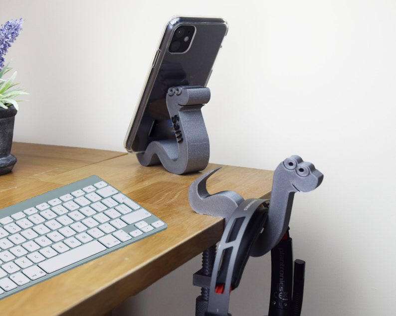 May include: Two 3D printed phone stands shaped like dinosaurs. The stand on the left is holding a black smartphone. The stand on the right is holding a pair of headphones. Both stands are on a wooden desk with a keyboard.