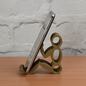 May include: A gold-colored phone stand shaped like a person sitting down. The stand is holding a silver smartphone.