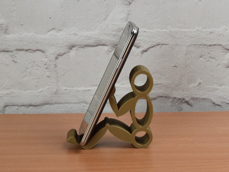 May include: A gold-colored phone stand shaped like a person sitting down. The stand is holding a silver smartphone.
