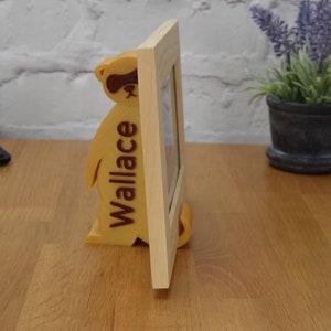May include: A wooden picture frame with a stand shaped like a raccoon. The raccoon is brown and has a black mask around its eyes. The frame is light brown and has the name "Wallace" engraved on the side.