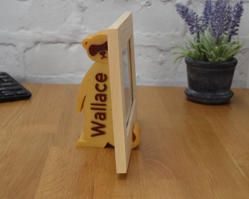 May include: A wooden picture frame with a stand shaped like a raccoon. The raccoon is brown and has a black mask around its eyes. The frame is light brown and has the name "Wallace" engraved on the side.