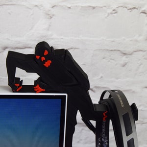 May include: A black 3D printed figure of a monster with red eyes and mouth, perched on top of a computer monitor. The figure is holding a pair of black headphones.
