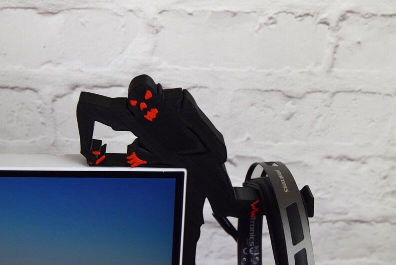 May include: A black 3D printed figure of a monster with red eyes and mouth, perched on top of a computer monitor. The figure is holding a pair of black headphones.