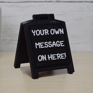 May include: A black A-frame sign with white lettering that reads "YOUR OWN MESSAGE ON HERE!". The sign is made of a matte black material and is designed to stand on a flat surface. The sign is a great way to display a custom message.