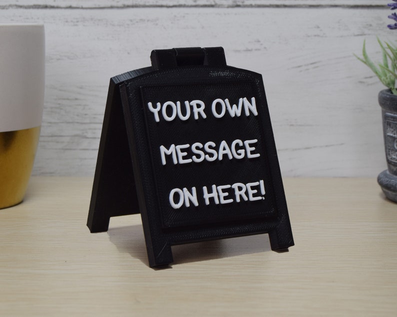 May include: A black A-frame sign with white lettering that reads "YOUR OWN MESSAGE ON HERE!". The sign is made of a matte black material and is designed to stand on a flat surface. The sign is a great way to display a custom message.