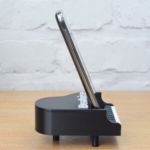 Puede incluir: Soporte para tel&eacute;fono negro con forma de piano con un tel&eacute;fono inteligente en su interior. El soporte tiene la palabra "Practice" impresa.