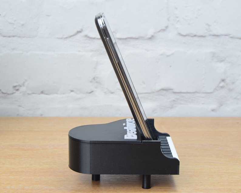 Puede incluir: Soporte para tel&eacute;fono negro con forma de piano con un tel&eacute;fono inteligente en su interior. El soporte tiene la palabra "Practice" impresa.