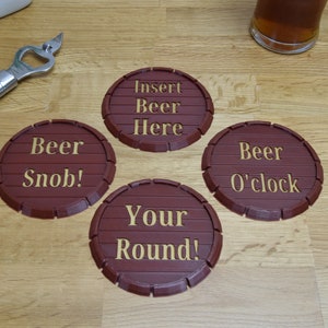 May include: Four brown, barrel-shaped coasters with gold lettering. The coasters read "Insert Beer Here," "Beer Snob!," "Beer O'clock," and "Your Round!" A silver bottle opener and a glass of beer are also visible.