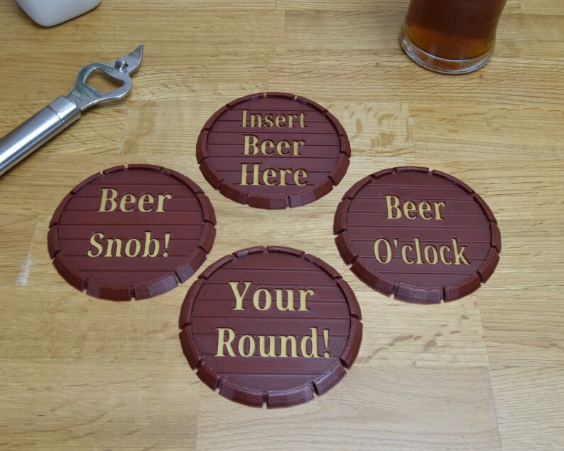 May include: Four brown, barrel-shaped coasters with gold lettering. The coasters read "Insert Beer Here," "Beer Snob!," "Beer O'clock," and "Your Round!" A silver bottle opener and a glass of beer are also visible.