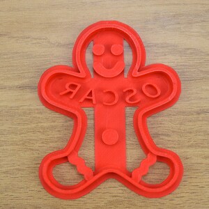 Puede incluir: Cortador de galletas de jengibre de pl&aacute;stico rojo en forma de hombre con la palabra "OSCAR" escrita en la parte delantera.